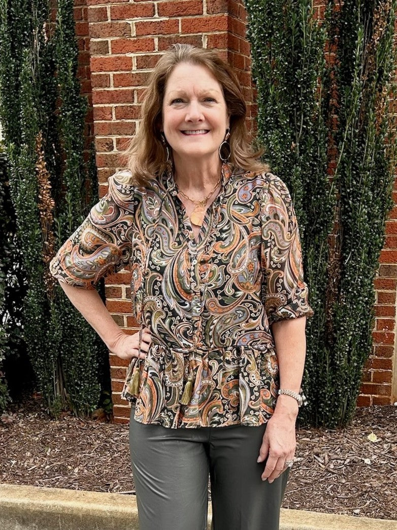 This lined paisley print top showcases beautiful fall colors, perfect for seasonal style. It features 3/4 length sleeves and a button-up front with a flattering V-neckline. The tassel tie adds a delicate detail, while the wide ruffled hem brings a feminine, flowing silhouette. This top combines comfort and elegance effortlessly.

LuAnn is wearing a size Small.

Material: 70% Rayon / 30% Nylon; Lining: 100% Polyester

Care Instructions: Hand wash cold, hang to dry