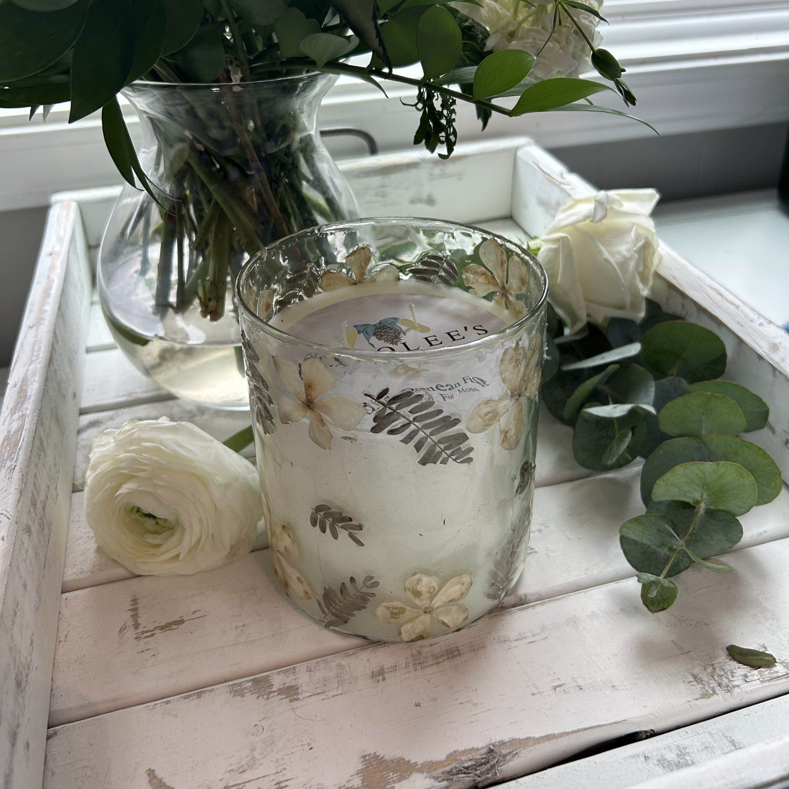 Illuminate your space with this elegant 18 oz candle featuring delicate flower petals and ferns suspended in a clear container. The medium-sized design fits perfectly on shelves, nightstands, or tables. Three wicks provide even, consistent burning and a beautiful ambient glow. Perfect for creating a serene atmosphere or as a thoughtful gift for nature lovers.

Scent: Mediterranean Fig (Green Leaves, Fig, Moss)