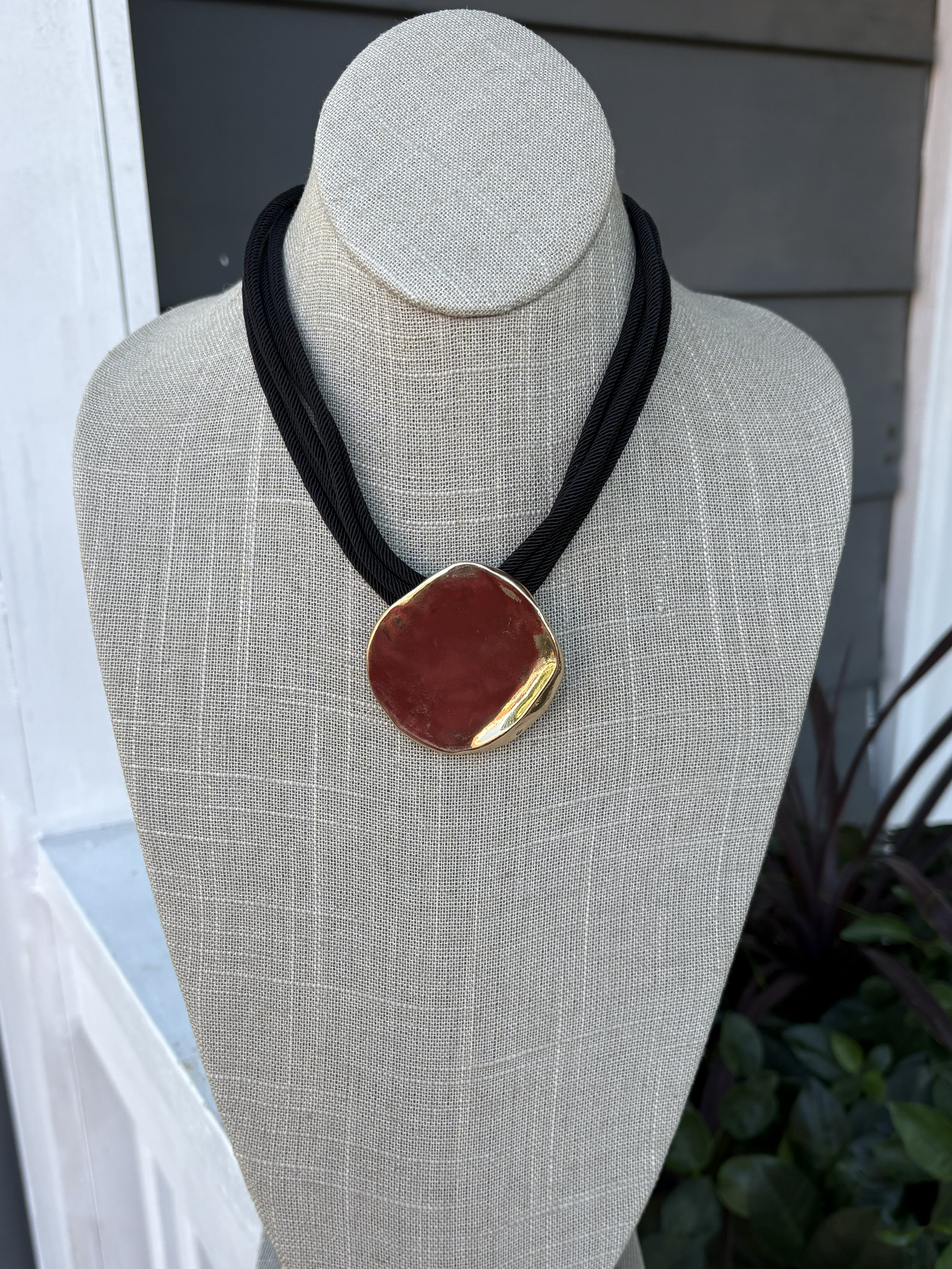 This is a striking black necklace featuring a gold hammered pendant and distinctive multi-rope chain design. Perfect for game day or any occasion where you want to make a bold statement. The textured pendant adds dimension and elegance to any outfit.