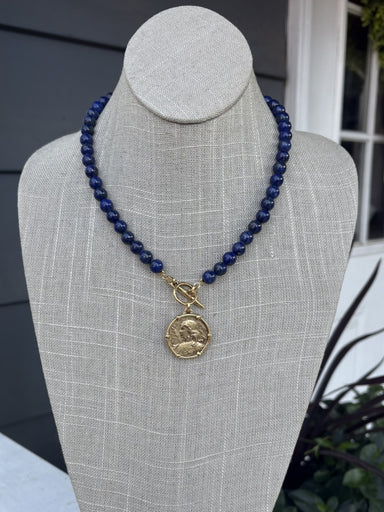 This dark blue beaded necklace showcases a captivating coin pendant that brings depth and character to any outfit. Each bead is carefully hand-strung to create a refined, textured look that works beautifully on its own or layered with other pieces. The secure T-bar closure provides a comfortable, reliable fit for all-day wear. Whether you're dressing up for an evening out or adding polish to casual wear, this versatile necklace effortlessly bridges your day and night wardrobe.