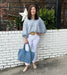 Tara is wearing a light blue and white checked blouse, white Risen jeans, and beige shoes, while holding a large textured blue tote bag.