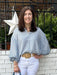 Tara is wearing a light blue and white checked blouse, white Risen jeans, and beige shoes, while holding a large textured blue tote bag.