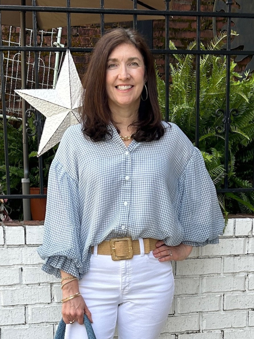 Tara is wearing a light blue and white checked blouse, white Risen jeans, and beige shoes, while holding a large textured blue tote bag.