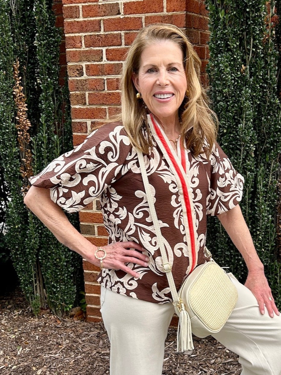 This Short Sleeve Ornate Design Top showcases a beautiful brown and beige print, perfectly capturing the warmth of fall colors. Its contrasting velour V-neckline adds a rich texture, while the short puffy sleeves with elastic closures create a flattering silhouette. Ideal as a stylish transition piece, this top effortlessly blends comfort and elegance for your seasonal wardrobe.

Barbara is wearing a size XSmall. She paired the top with our "Erin Wide Leg Denim/Pants in Oat. She added our "Marla Leather Raf