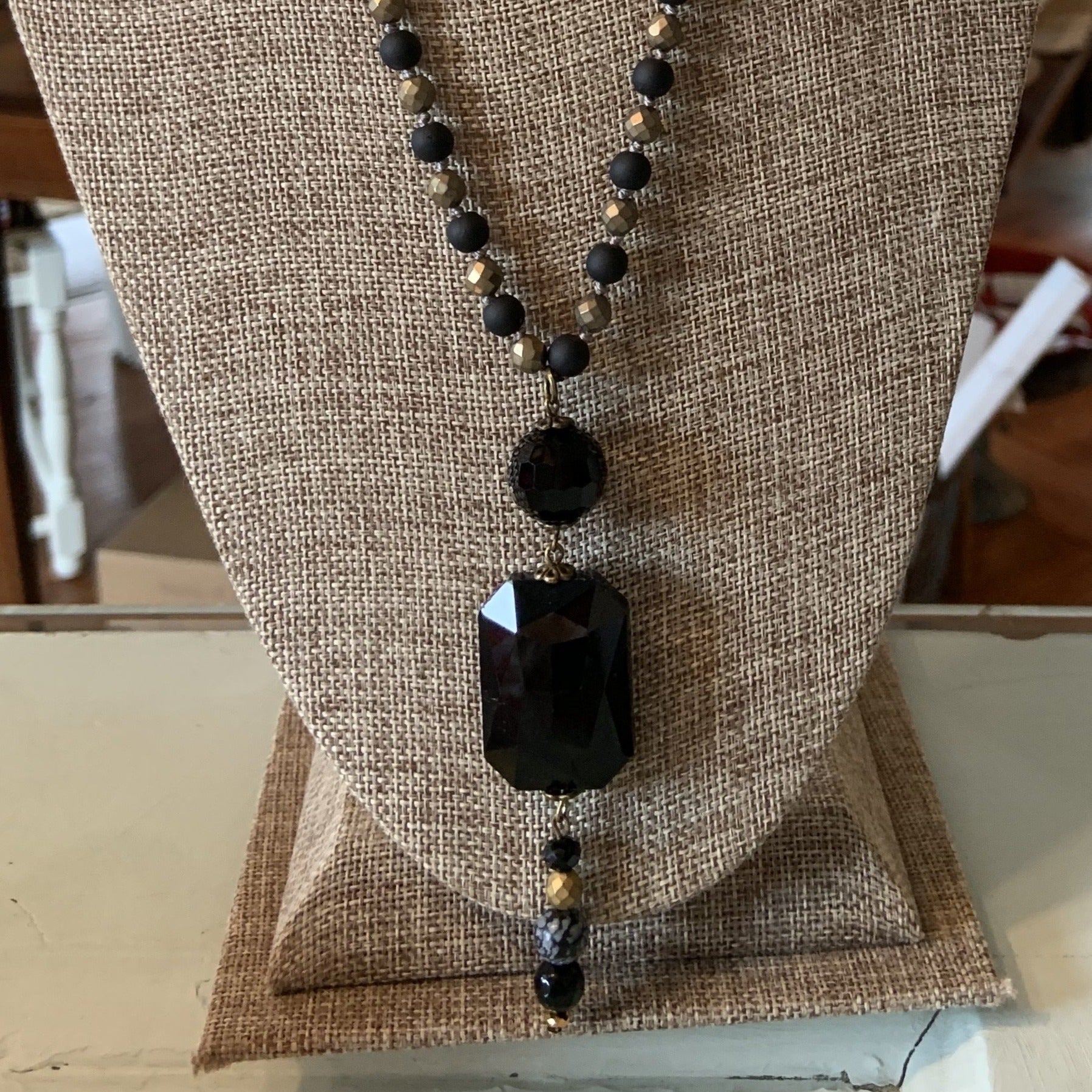 Black and gold long necklace with stacked small black circle, large black rectangle finally followed by 4 smaller black, gold and grey beads